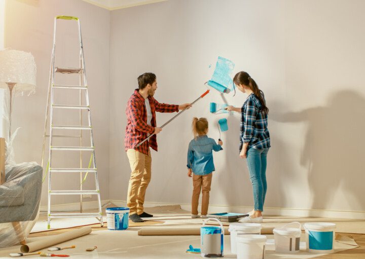 Foto de uma mulher, um homem e uma criança pequena pintando a parede de casa com um rolo pintura mergulhado em tinta azul para ilustrar matéria sobre como economizar na reforma da casa