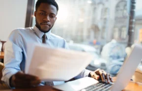 Um homem negro, vestido de roupa social, analisa uma documentação com auxílio do notebook.