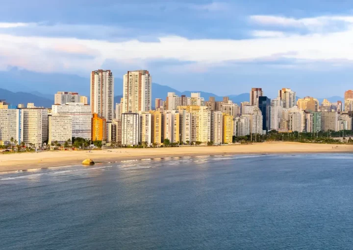 Foto que ilustra matéria sobre o que fazer em São Vicente mostra uma vista panorâmica da faixa de areia da praia de Itararé, com o mar em primeiro plano e prédios altos ao fundo