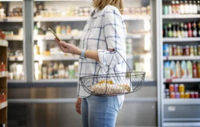 Imagem de uma mulher branca vestindo calça jeans e camiseta listrada, com uma cesta de compras pendurada no braço, parada em frente a uma prateleira de produtos para ilustrar matéria sobre mercado em condomínio