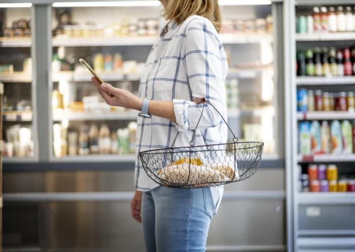 Imagem de uma mulher branca vestindo calça jeans e camiseta listrada, com uma cesta de compras pendurada no braço, parada em frente a uma prateleira de produtos para ilustrar matéria sobre mercado em condomínio