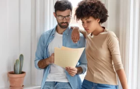 Imagem de um casal composto por um homem e uma mulher um ao lado do outro concentrados em ler as informações descritas em um papel para ilustrar matéria sobre multa por quebra de contrato por parte do locador