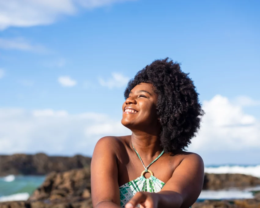 Imagem de uma mulher negra sorridente vestindo uma camisa azul sentada em uma praia olhando para o lado com um céu azul ao fundo para ilustrar matéria sobre as cidades mais seguras da Bahia