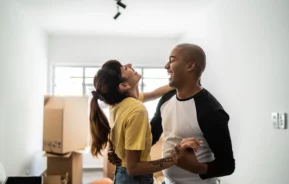 Imagem de um casal composto por um homem e uma mulher dançando com expressão de felicidade no rosto em sua casa nova para ilustrar matéria sobre como financiar 100% de um imóvel