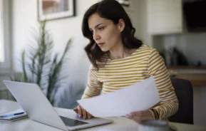 Imagem de uma mulher jovem e branca com uma expressão séria sentada olhando para a tela de um computador com alguns papéis na mão para ilustrar a consulta de imóveis por CPF grátis