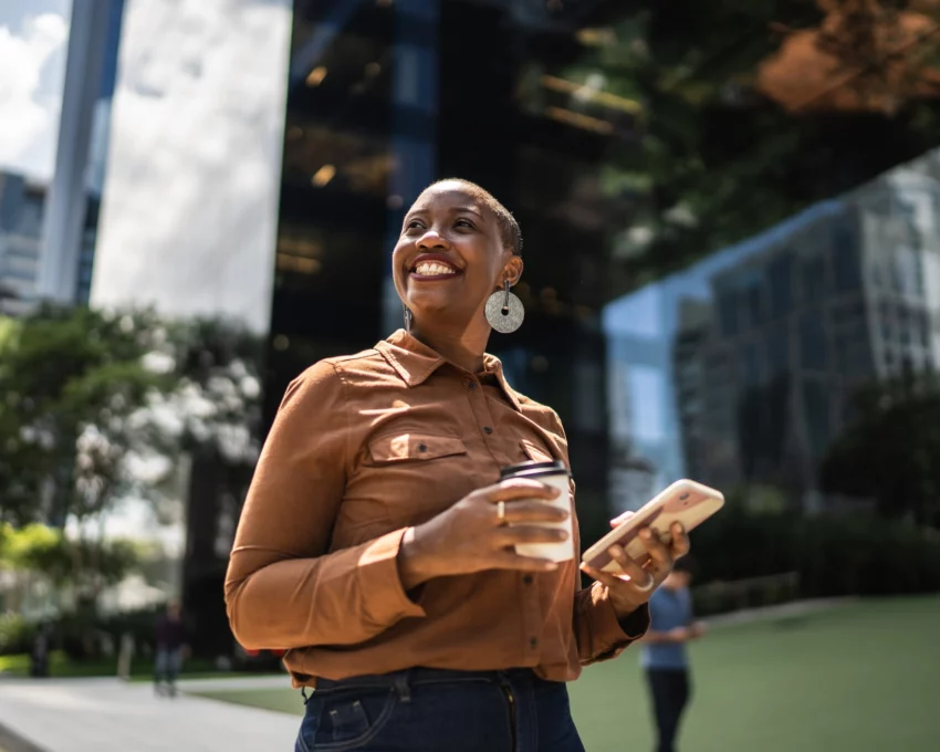 Imagem de uma mulher negra andando em uma rua em frente a um prédio empresarial, sorrindo e com um celular na mão, para ilustrar matéria sobre qual a cidade mais tranquila do Brasil