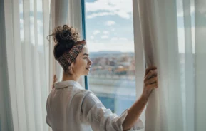 Imagem de uma mulher jovem e sorridente abrindo as cortinas de um apartamento e olhando para o horizonte para ilustrar matéria sobre quanto custa um apartamento