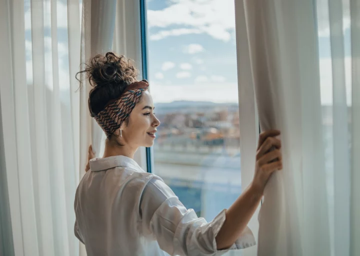 Imagem de uma mulher jovem e sorridente abrindo as cortinas de um apartamento e olhando para o horizonte para ilustrar matéria sobre quanto custa um apartamento