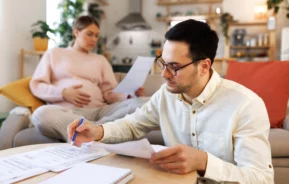 Imagem de uma mulher grávida sentada em um sofá enquanto um homem olha para alguns papéis em cima de uma mesa para ilustrar matéria sobre a seguinte dúvida: sou casado, posso financiar um imóvel sozinho?