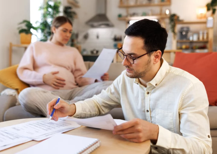 Imagem de uma mulher grávida sentada em um sofá enquanto um homem olha para alguns papéis em cima de uma mesa para ilustrar matéria sobre a seguinte dúvida: sou casado, posso financiar um imóvel sozinho?