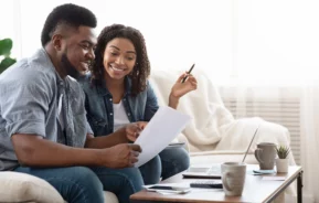 Imagem de um casal de jovens negros composto por um homem e uma mulher sorridentes sentados no sofá de uma casa olhando para um documento para ilustrar matéria sobre termo de quitação de imóvel