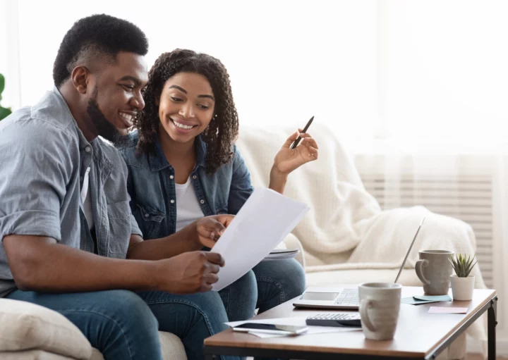 Imagem de um casal de jovens negros composto por um homem e uma mulher sorridentes sentados no sofá de uma casa olhando para um documento para ilustrar matéria sobre termo de quitação de imóvel