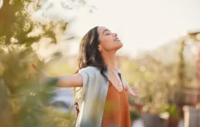 Foto de uma mulher sorridente com os braços abertos, em meio à natureza.