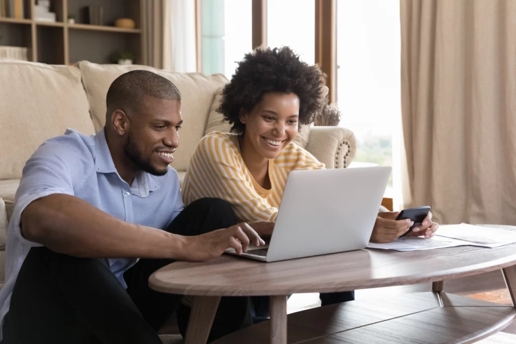 Imagem e um casal composto por um homem e uma mulher negros sentados no chão de uma sala olhando para o computador em cima de uma mesa de centro para ilustrar matérias sobre as possibilidades disponíveis na hora de saber como alugar imóvel com score baixo
