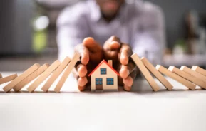 Imagem de um dominó caindo sobre a miniatura de uma casa e sendo barrado pelas mãos de uma pessoa para ilustrar matéria sobre seguro prestamista