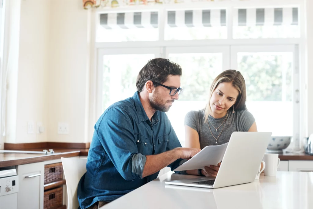 Imagem de um casal composto por um homem e uma mulher sentados em uma mesa da cozinha analisando informações no computador em cima da mesa para ilustrar matéria sobre as coberturas do seguro prestamista