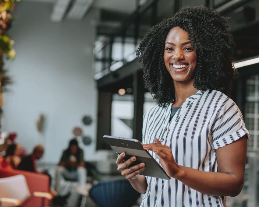 Mulher negra em ambiente empresarial sorrindo e segurando um tablet para ilustrar matéria sobre as cidades com mais empregos no Brasil