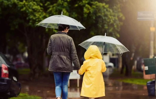 Imagem de uma mulher e uma criança de costas andando em uma faixa de pedestres com guarda chuva na mão para ilustrar matéria sobre as cidades que mais chovem no Brasil