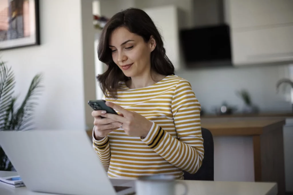 Imagem de uma mulher jovem sentada em frente a um computador e mexendo no celular para ilustrar matéria sobre qual score ideal para financiamento de imóvel