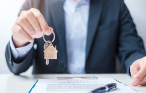 Imagem de um homem de terno segurando uma chave com chaveiro de casa enquanto apresenta uma prancheta com documento e caneta para ilustrar matéria sobre despachante imobiliário