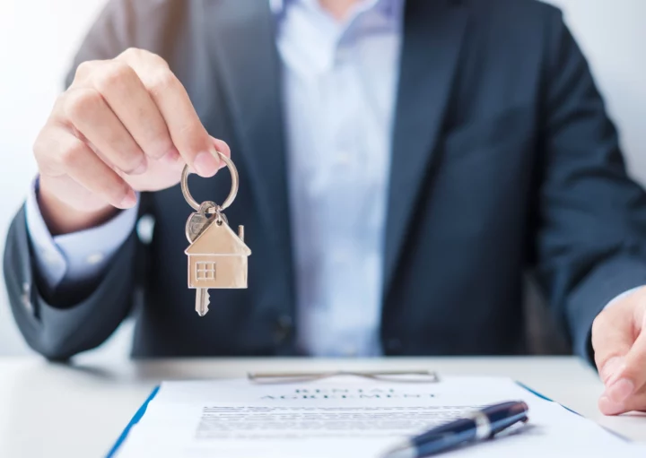 Imagem de um homem de terno segurando uma chave com chaveiro de casa enquanto apresenta uma prancheta com documento e caneta para ilustrar matéria sobre despachante imobiliário