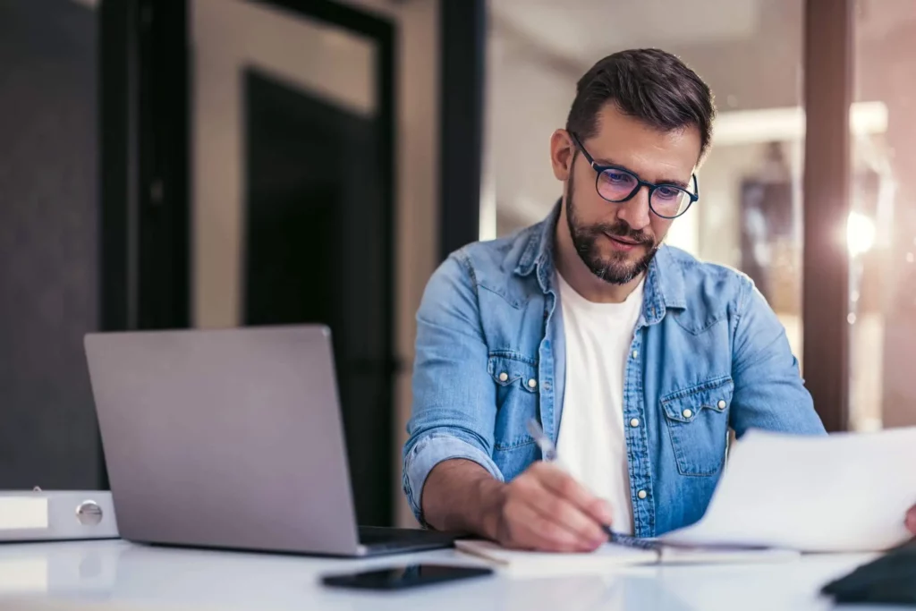 Imagem de um homem branco de óculos e barba usando camiseta jeans escrevendo em um papel ao lado de um computador que está em cima de uma mesa para ilustrar matéria sobre o prazo para informar reajuste de aluguel
