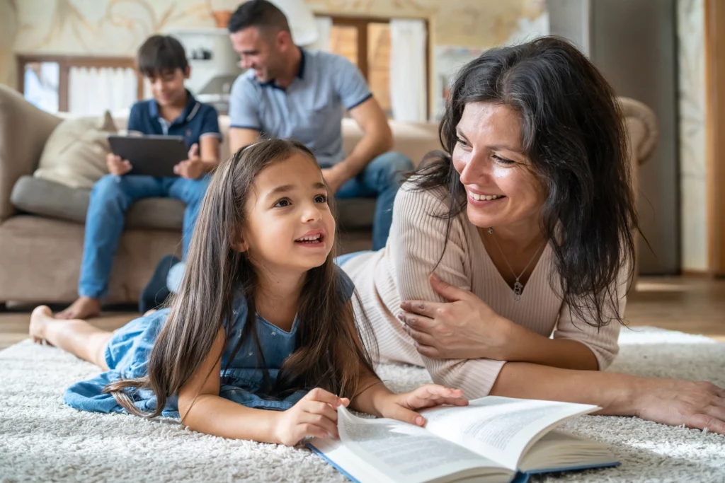 Imagem de uma família na sala de casa onde a mãe está deitada no chão com a filha lendo um livro e o pai está sentado no sofá com o filho olhando o tablet para ilustrar matéria sobre qual o tamanho ideal de um apartamento para 4 pessoas
