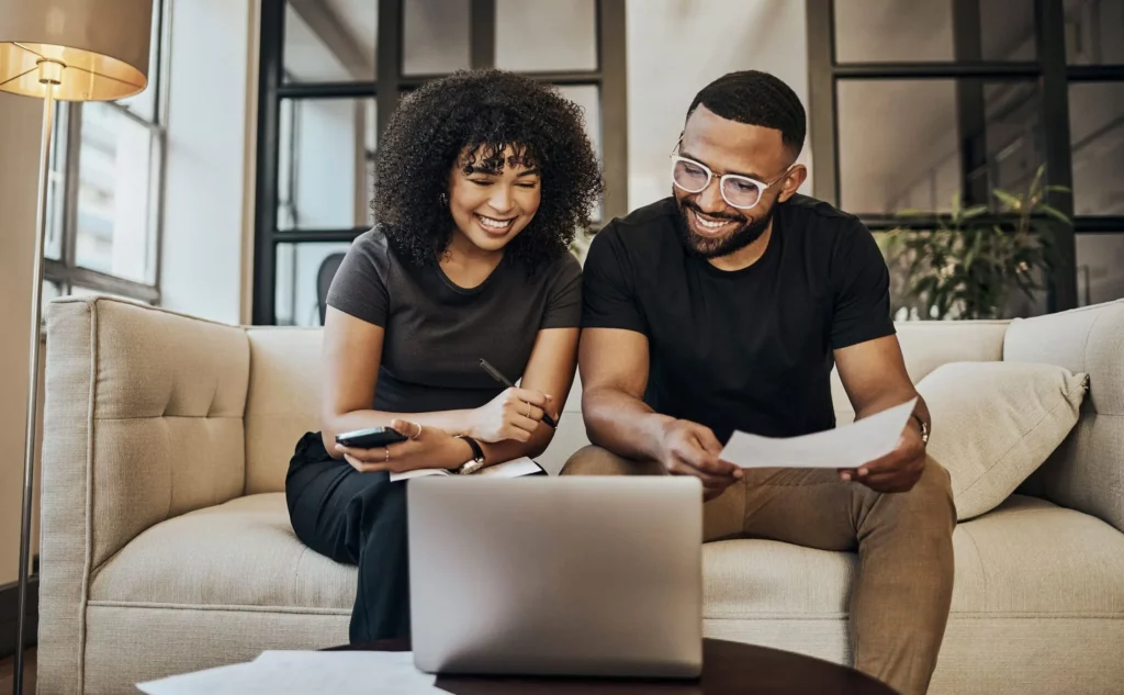 Imagem de uma mulher e de um homem sorrindo, sentados no sofá em frente a um computador, para ilustrar matéria sobre qual score ideal para financiamento de imovel