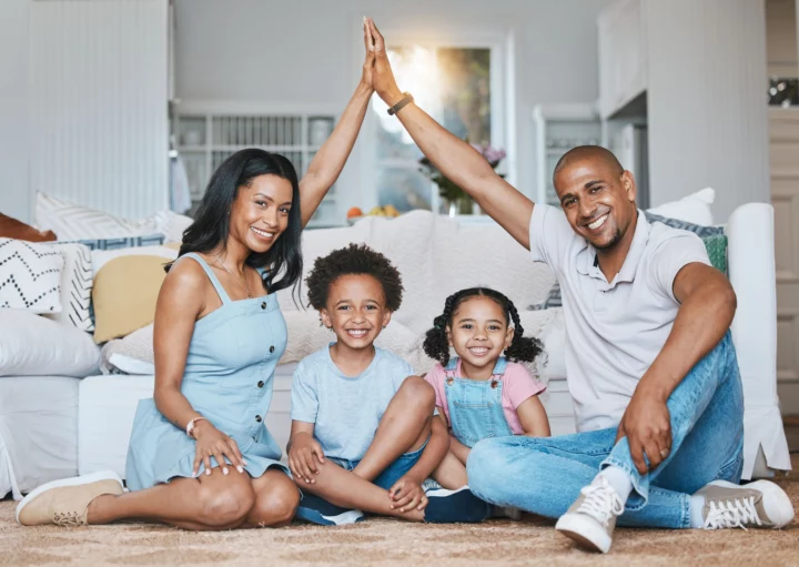 Imagem de uma família composta por um casal e duas crianças negras sentadas no chão da sala sorridentes para ilustrar matéria sobre o tamanho ideal de apartamento para 4 pessoas