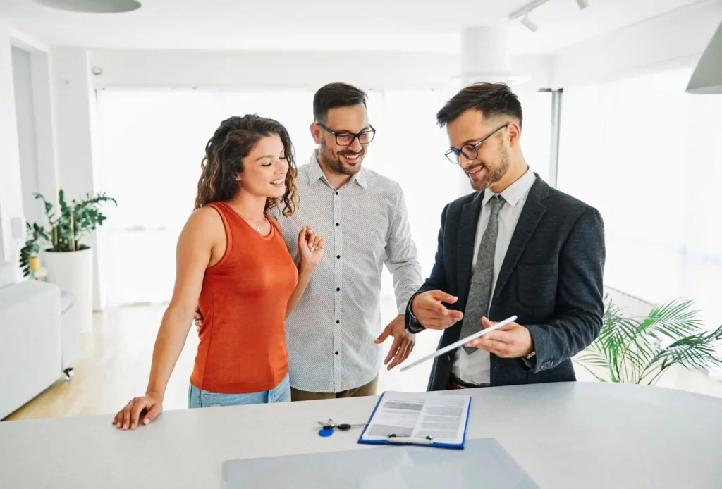 Imagem de um casal em um imóvel vazio conversando com um profissional imobiliário de terno para ilustrar a matéria sobre assinatura de contrato de locação