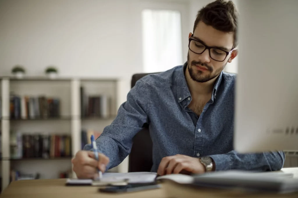 Imagem de um homem sentado na frente do computador escrevendo em um caderno para ilustrar matéria sobre como escrever endereço de apartamento