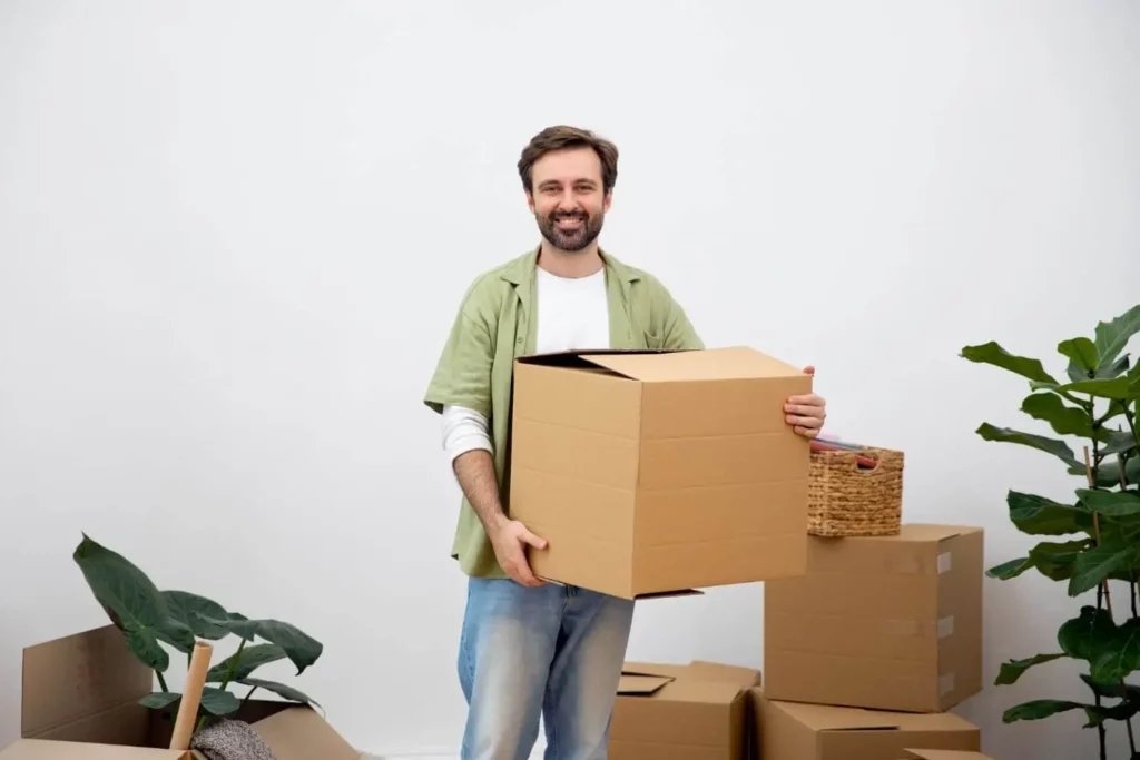 Imagem de um homem jovem segurando uma caixa de mudança na mão em um cômodo sem móveis e com mais caixas de mudança para ilustrar matéria sobre documentos para contrato de aluguel