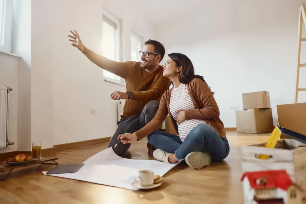 Imagem de um casal composto por um homem e uma mulher sentados no chão de um imóvel com alguns papéis e caixas de papelão à vista para ilustrar matéria sobre fazer financiamento de imóvel no nome de outra pessoa