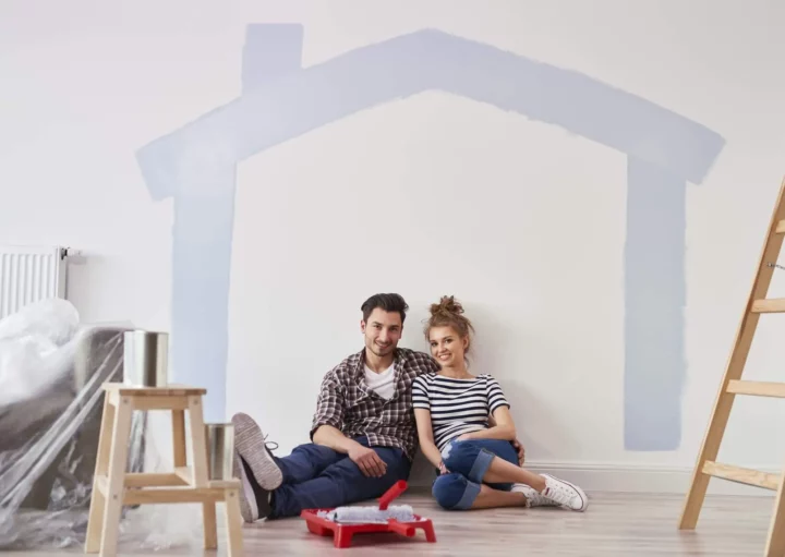 Imagem de um casal composto por um homem e uma mulher sentados no chão de uma casa com alguns itens de obra ao lado e o desenho de uma casa na parede branca para ilustrar matéria sobre habitação de interesse social