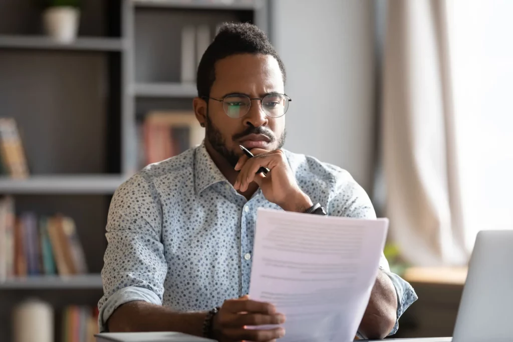 Imagem de um homem sentado em frente a um computador, lendo um papel atentamente para ilustrar a matéria sobre o que o que conferir no modelo de contrato de locação