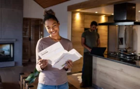 Imagem de uma mulher dentro de casa olhando para uma conta que chegou pelos Correios enquanto o marido está atrás mexendo no computador para ilustrar matéria sobre o que é código postal