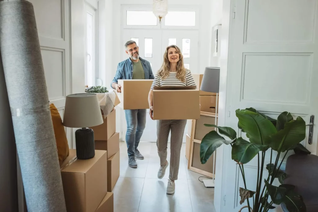 Imagem de um casal entrando em uma casa segurando caixas de papelão para ilustrar matéria sobre o que fazer quando o IPTU está no nome de outra pessoa