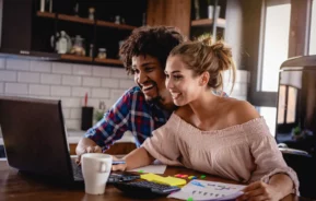 Imagem de um casal composto por um homem e uma mulher olhando para a tela de um computador sentados na mesa de uma cozinha para ilustrar matéria sobre paga