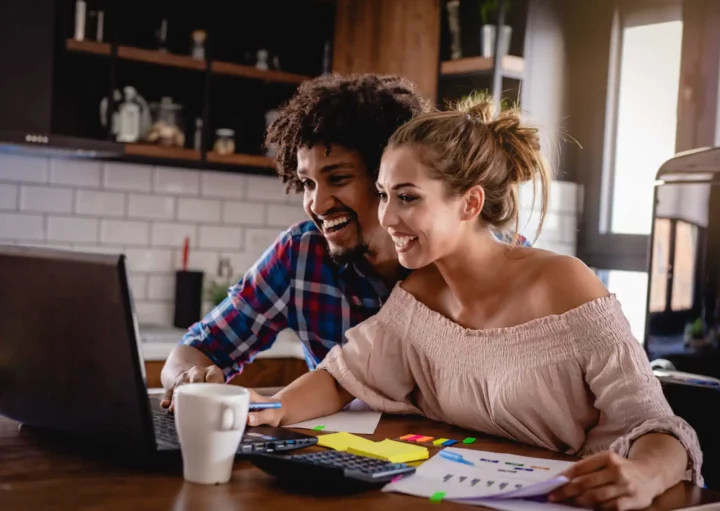 Imagem de um casal composto por um homem e uma mulher olhando para a tela de um computador sentados na mesa de uma cozinha para ilustrar matéria sobre paga