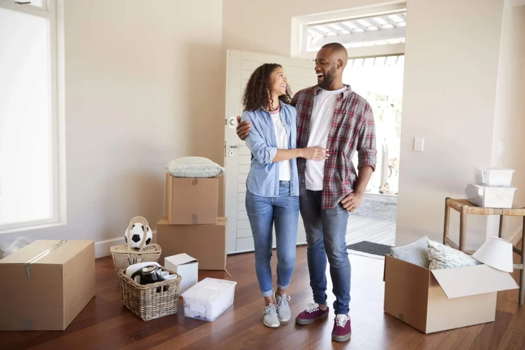 Imagem de um casal entrando em uma casa nova com algumas caixas de papelão espalhadas pela sala para ilustrar matéria sobre programa habitacional em SP
