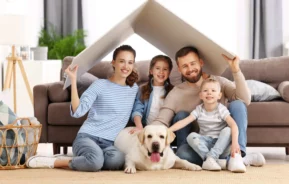 Imagem de uma família composta por um pai, uma mãe, dois filhos crianças e um cachorro sentados no chão da sala com um papelão em cima da cabeça sinalizando o telhado de uma casa para ilustrar matéria sobre como aumentar a segurança em apartamento