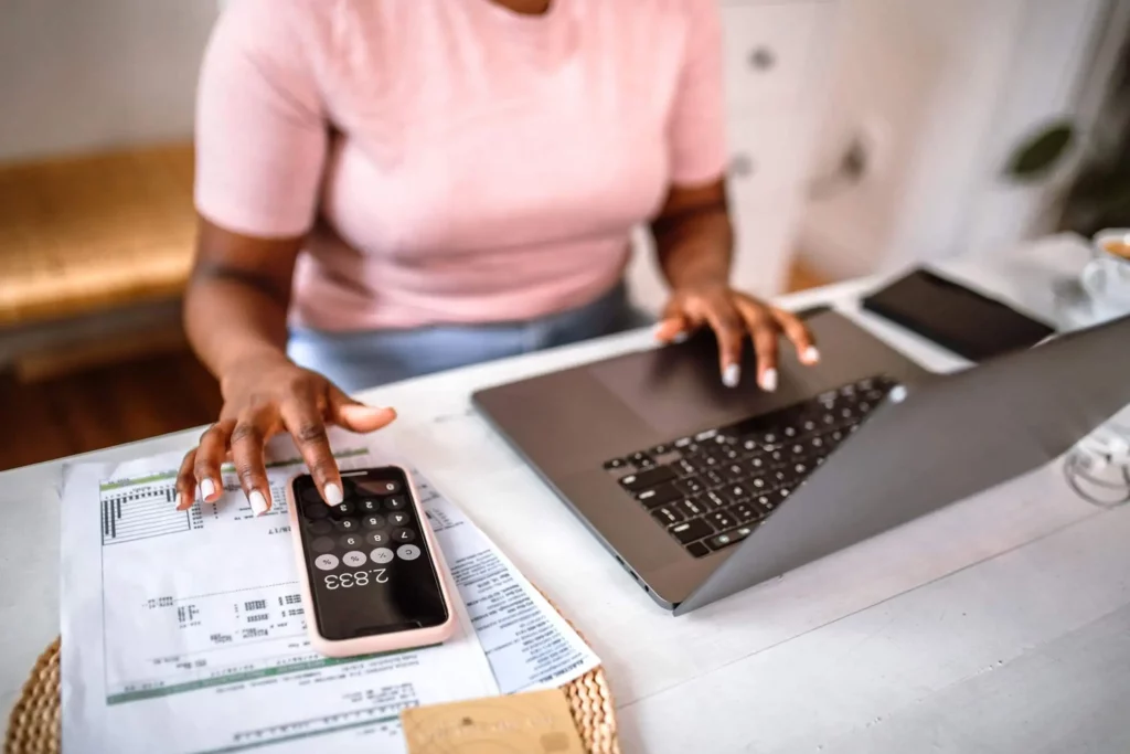 Imagem de uma mulher fazendo as contas de casa na calculadora de um celular que está em cima de uma mesa ao lado de contas e um computador para ilustrar matéria sobre como calcular IGP-M negativo no aluguel