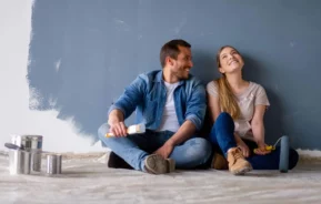 Imagem de um casal composto por um homem e uma mulher sentados no chão de um imóvel ao lado de latas de tinta enquanto ao fundo a parede está quase toda pintada na cor azul para ilustrar matéria sobre construir ou reformar