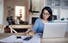 Imagem de uma mulher sentada na mesa de casa com um computador aberto, papéis ao lado e fazendo contas no celular para ilustrar matéria para responder à dúvida se desempregado pode financiar imóvel pela Caixa