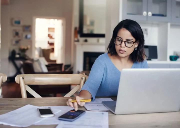 Imagem de uma mulher sentada na mesa de casa com um computador aberto, papéis ao lado e fazendo contas no celular para ilustrar matéria para responder à dúvida se desempregado pode financiar imóvel pela Caixa