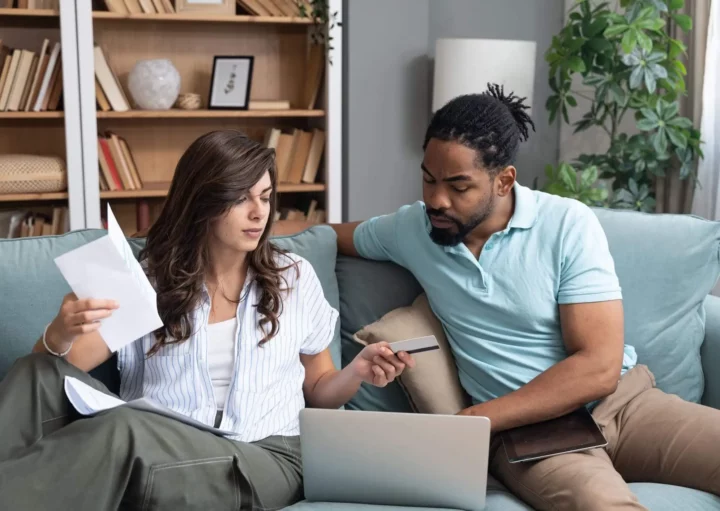 Imagem de um casal composto por um homem e uma mulher sentados no sofá de casa fazendo contas com um computador entre eles para ilustrar matéria sobre o que é IGP-M