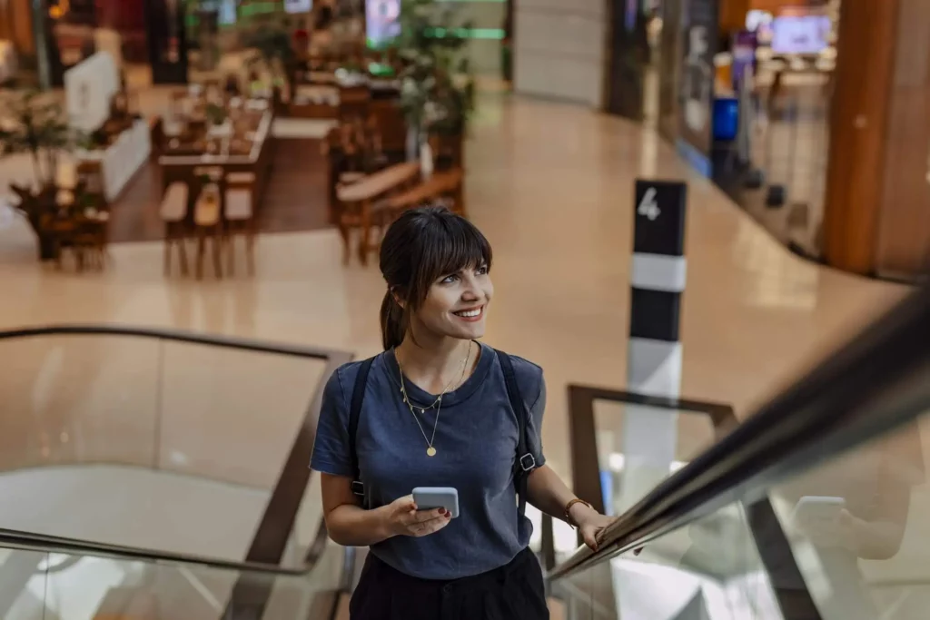 Imagem de uma jovem mulher sorrindo com o celular na mão enquanto usa a escada rolante de um shopping center para ilustrar matéria sobre Shopping Villa-Lobos, que é acessível pelo metrô de São Paulo, via linha 9-Esmeralda