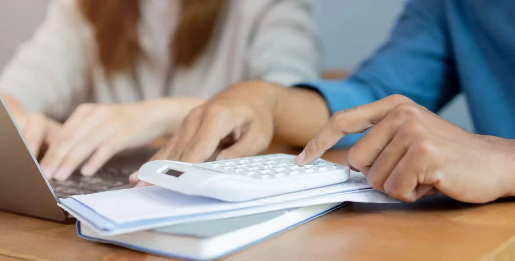 Imagem de duas pessoas fazendo contas em uma calculadora e em um notebook que estão em cima de uma mesa para ilustrar matéria sobre o valor do aluguel quando o IGP-M é negativo