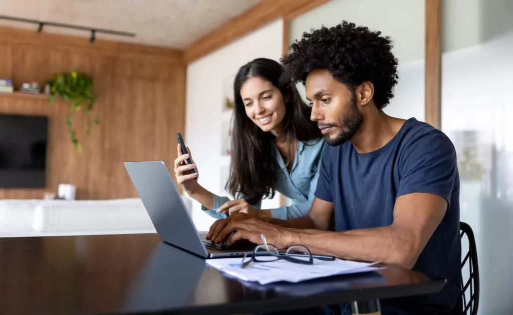 Imagem de um casal composto por um homem e uma mulher sentados na mesa da cozinha olhando para a tela de um computador para ilustrar matéria sobre o valor do IGP-M hoje