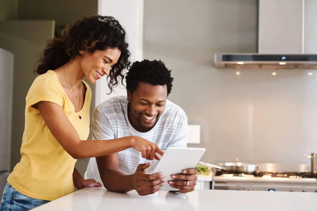 Imagem de um casal de homem e mulher encostados na bancada da cozinha olhando para um tablet para ilustrar matéria sobre cessão de imóvel.